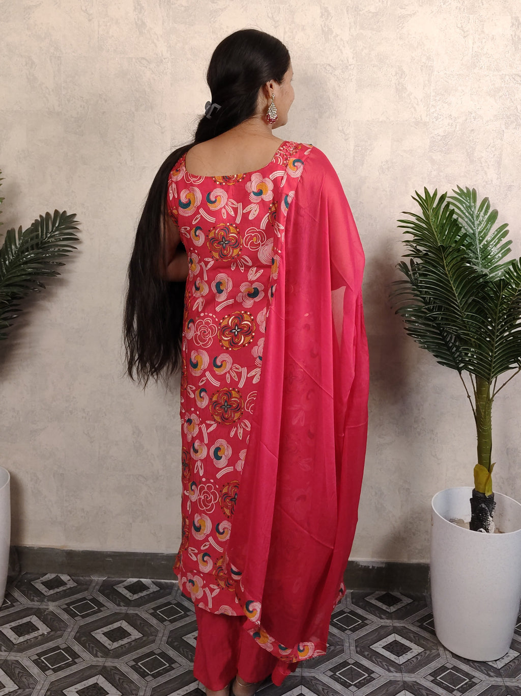 Woman in a pink saree with floral patterns standing against a light-colored wall.