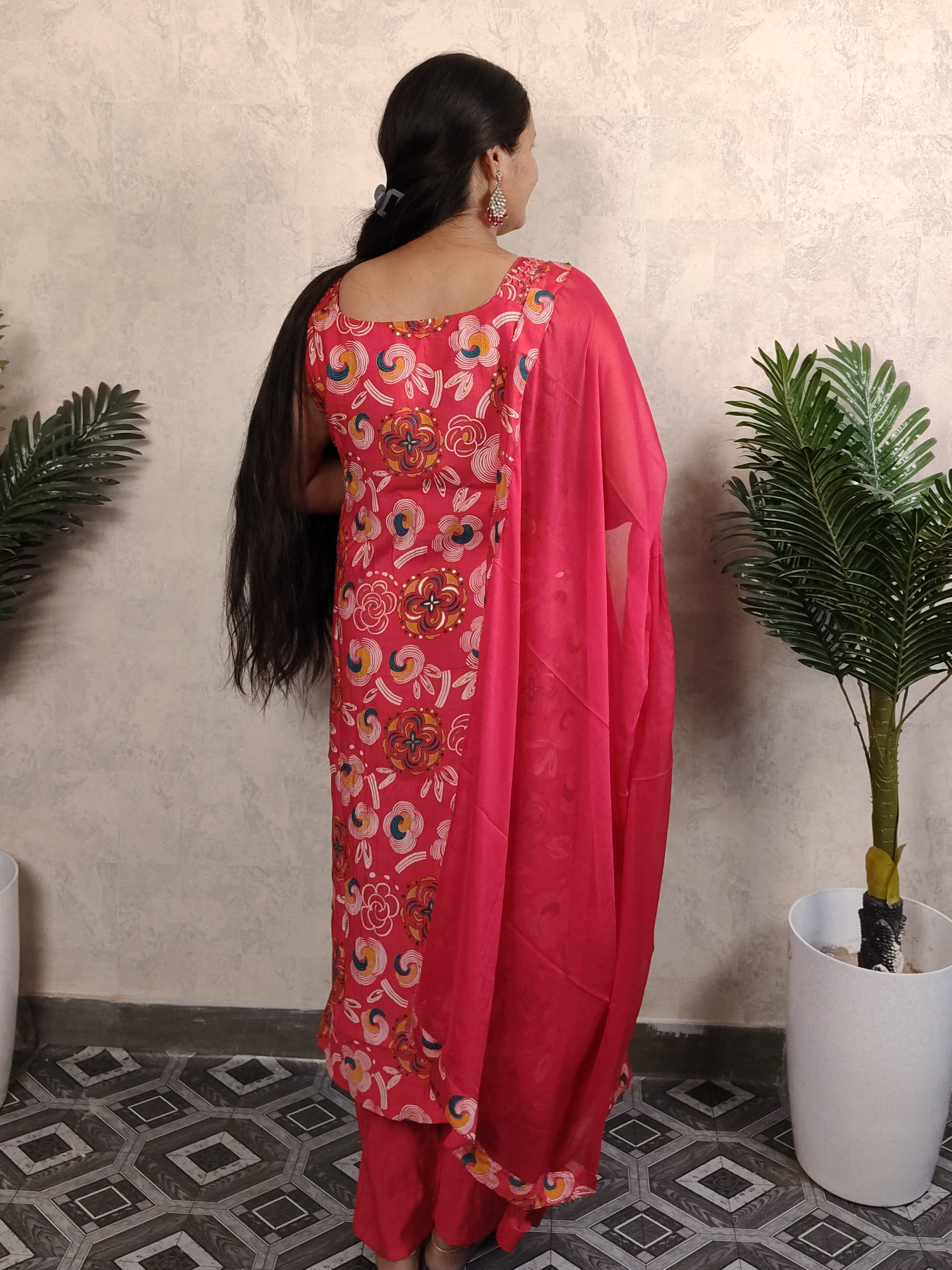 Woman in a pink saree with floral patterns standing against a light-colored wall.