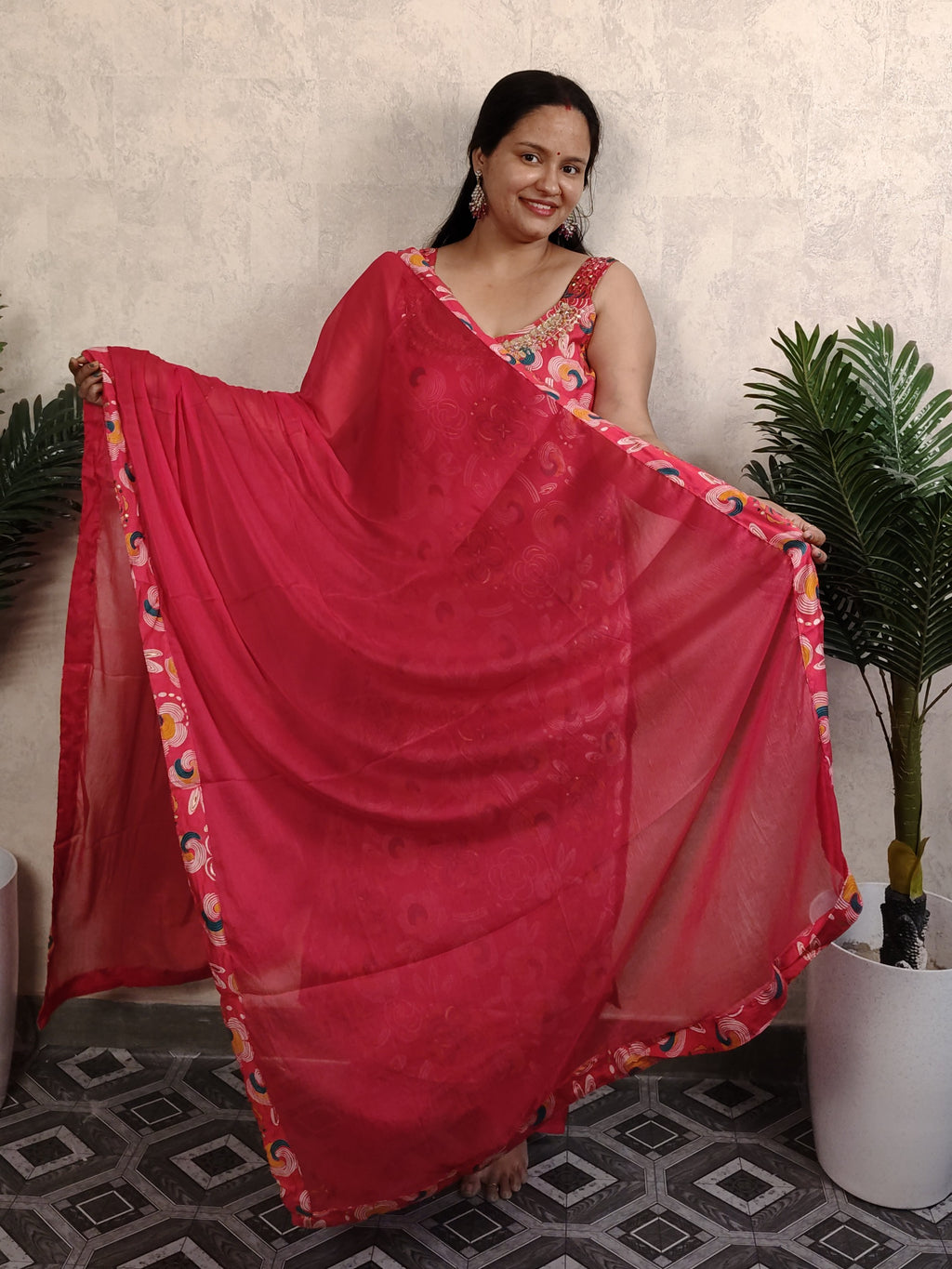 Woman holding a red saree with a floral border in a room with plants.