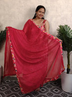 Woman holding a red saree with a floral border in a room with plants.