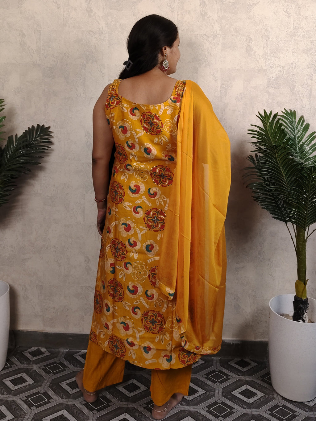 Woman in a yellow floral dress standing against a neutral wall with plants on either side.