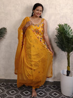 Woman in a yellow saree with floral patterns standing indoors next to a plant.